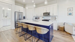 A modern kitchen with white cabinetry, a stainless steel refrigerator, a large island with bar stools, and pendant lighting. - Spacious & modern home with gleaming kitchen, pool access & balcony (Port Aransas)