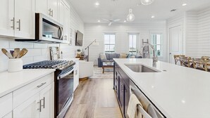 A modern kitchen with white cabinetry, a central island with a sink, and a dining area with a table and chairs. - Spacious & modern home with gleaming kitchen, pool access & balcony (Port Aransas)