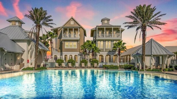 A large swimming pool surrounded by multi-story buildings with balconies, palm trees, and a sunset sky. - Spacious & modern home with gleaming kitchen, pool access & balcony (Port Aransas)