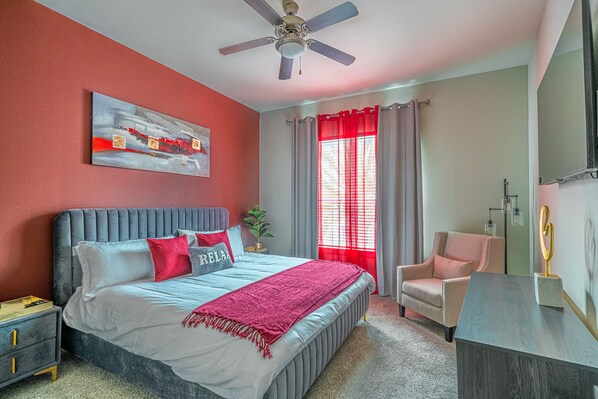 A bedroom with a bed, a chair, a desk, a ceiling fan, a window with curtains, and a painting on the wall.