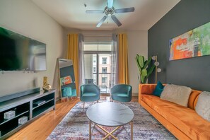 Un salon moderne avec un téléviseur à écran plat, deux fauteuils turquoise, une table ronde, un canapé orange, un ventilateur de plafond et une grande fenêtre ornée de rideaux jaunes.
