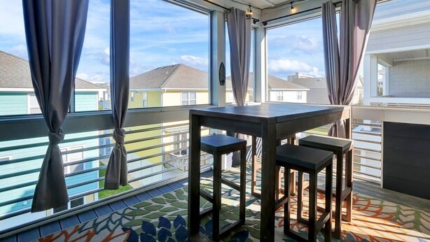 A balcony with a table and stools, large windows with curtains, and a view of neighboring houses. - SeaRenity with rooftop and ocean views (Carolina Beach)