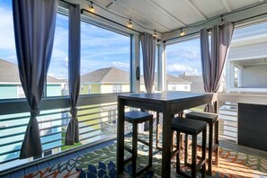 Een balkon met een tafel en krukjes, grote ramen met gordijnen, en uitzicht op omliggende huizen.