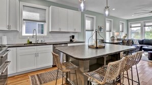 A modern kitchen with white cabinets, a central island with bar stools, pendant lighting, and a view of the living area with a sofa and coffee table. - SeaRenity with rooftop and ocean views (Carolina Beach)