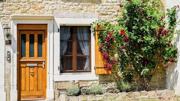 A stone building with a wooden door and window, number 107, and a vine with red flowers growing on the wall.