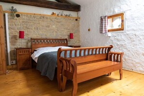Un dormitorio rústico con pared de piedra, cama de madera, banco y una ventana pequeña con cortinas.