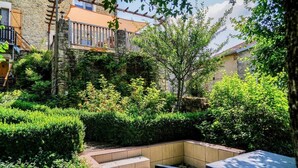 A stone building with a wooden balcony, a garden with hedges, and a seating area with steps.
