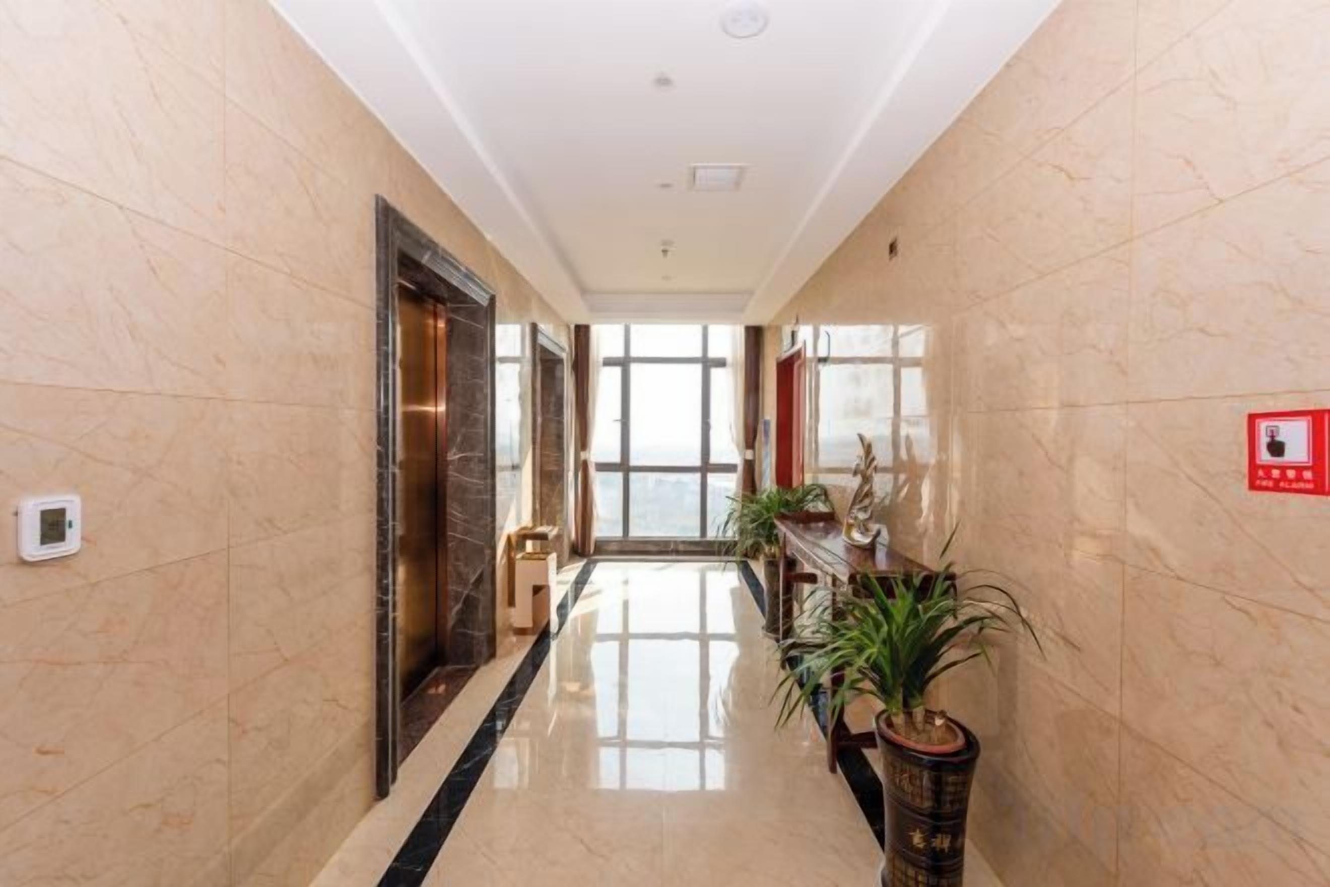 A hallway with a marble floor, a fire alarm on the wall, and a potted plant.