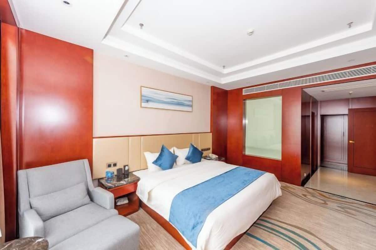 A hotel room with a large bed, a grey armchair, a bedside table with a lamp, and a window with a view.