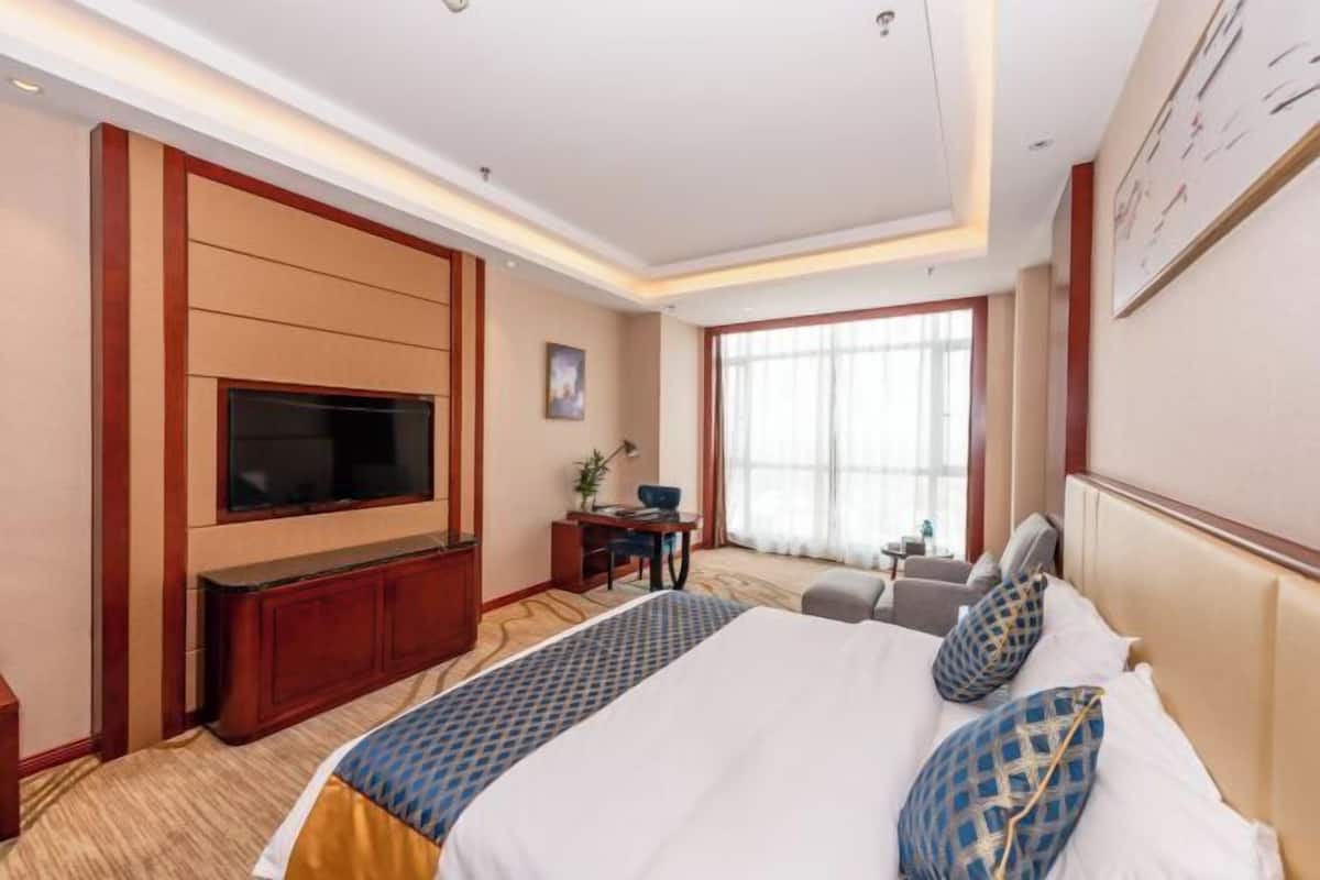 A hotel room with a large bed, a flat-screen TV mounted on the wall, a desk with a chair, and a window with curtains.