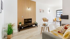 A modern living room with a TV, wooden panel wall, a dining area with a table and chairs, and a sofa with cushions. - Sunny Duplex W/ Pool & Balcony by LovelyStay (Carvoeiro)