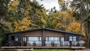 A modern cabin with a deck, large windows, and a forest backdrop. - Sea, Sunrises, Sauna | Oceanfront Group Retreat (Campbell River)