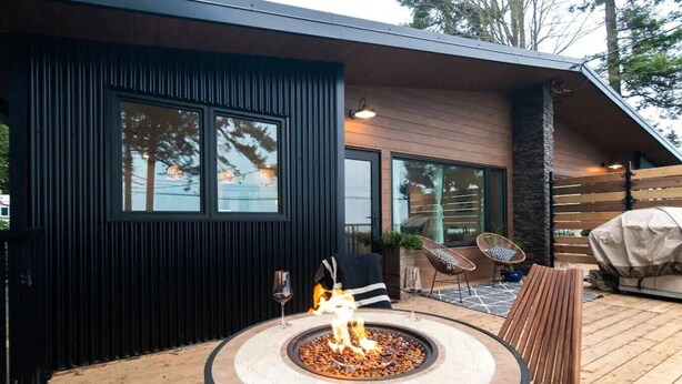 A modern outdoor patio with a circular fire pit, two wicker chairs, and a wooden bench. - Sea, Sunrises, Sauna | Oceanfront Group Retreat (Campbell River)