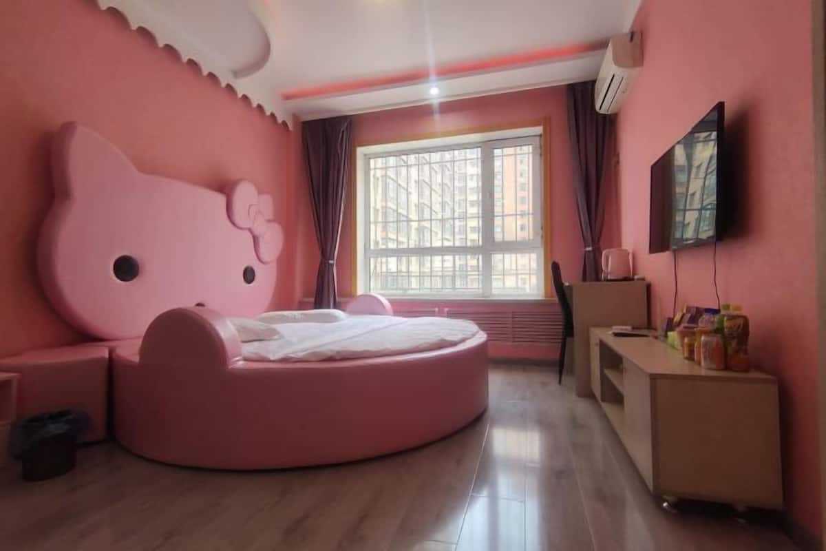 A pink-themed bedroom with a round bed, a TV mounted on the wall, and a window with curtains.