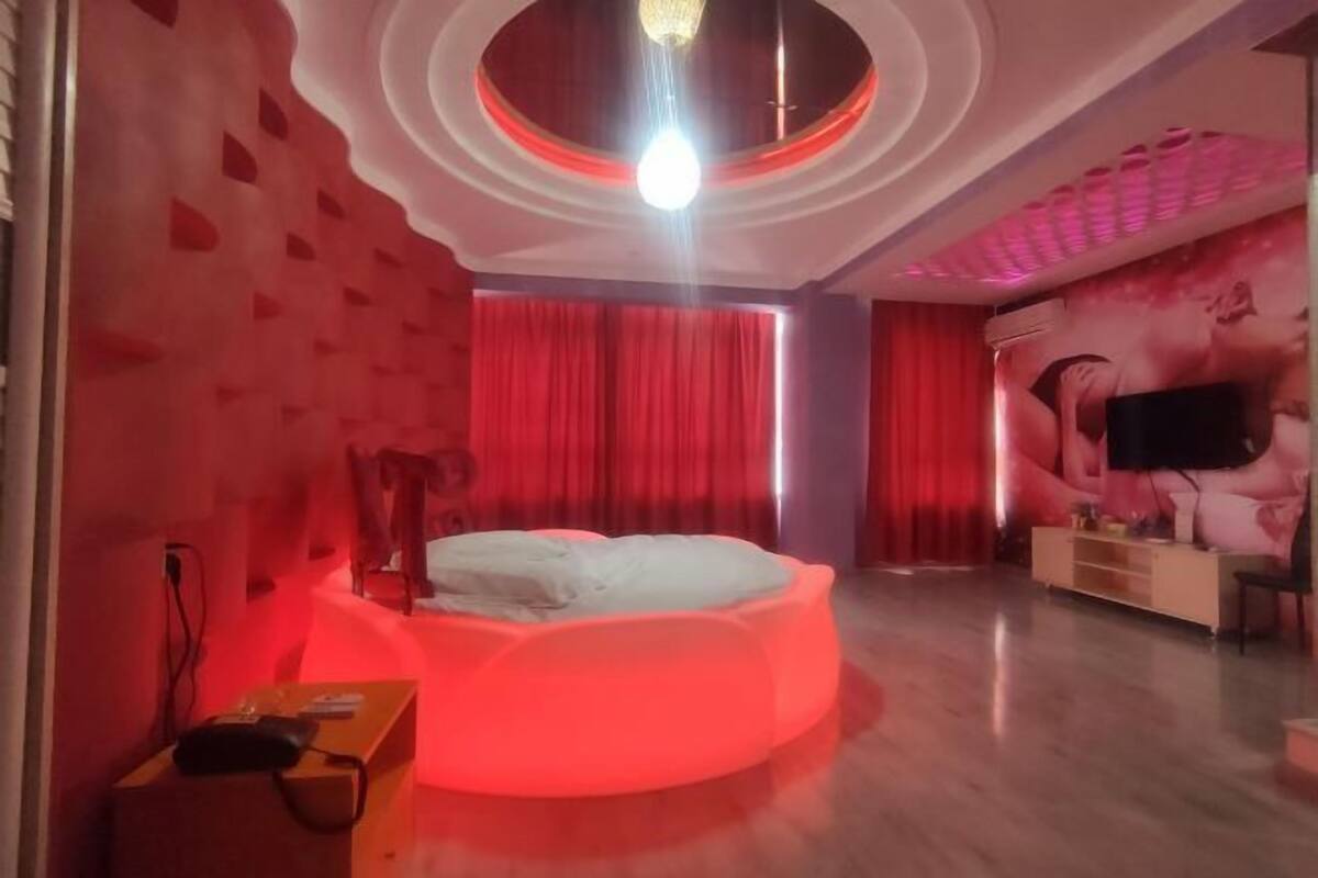 A room with a circular ceiling design, a bed with a red-lit base, a flat-screen TV, and a wall with a large poster.