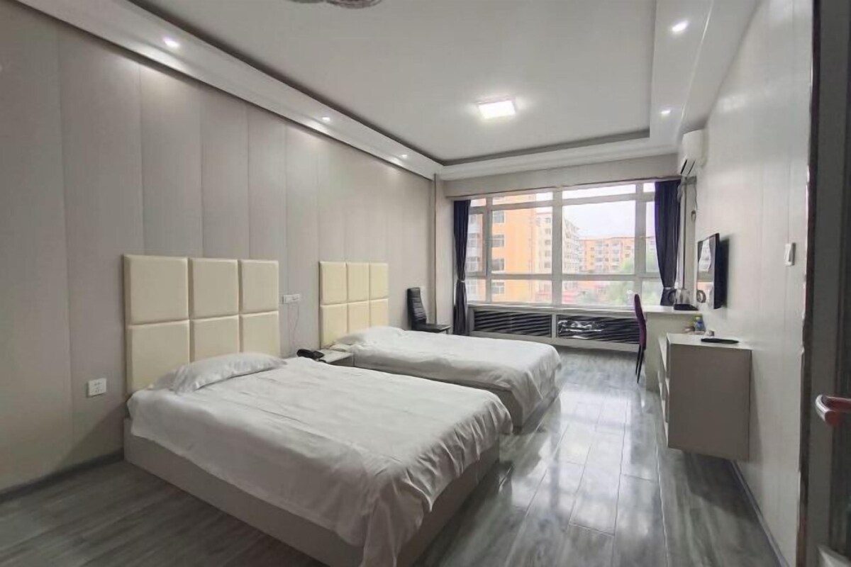 A hotel room with two beds, a desk, a chair, a TV, and a window with a view of buildings.