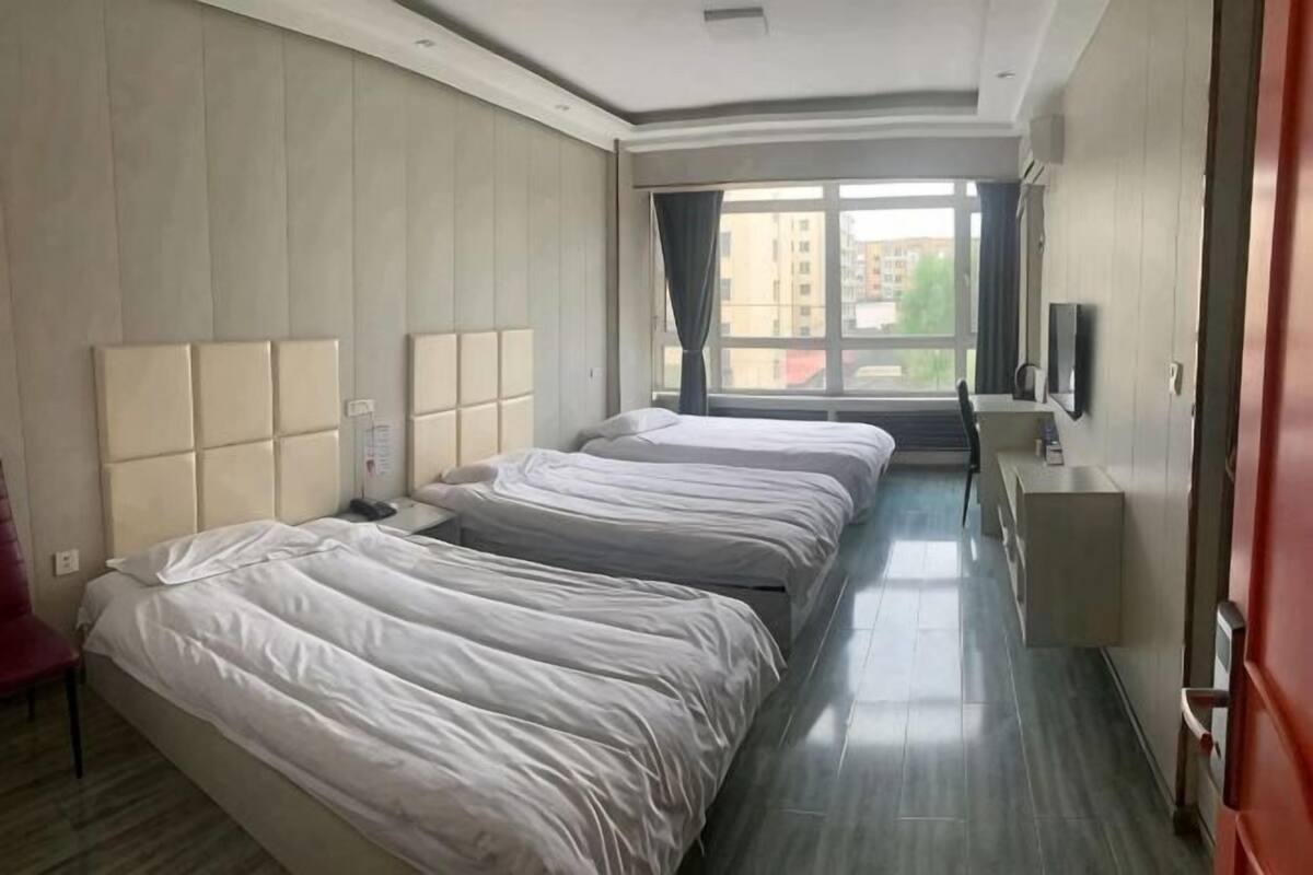 A hotel room with three beds, a TV, a desk, and a window with a view of buildings.