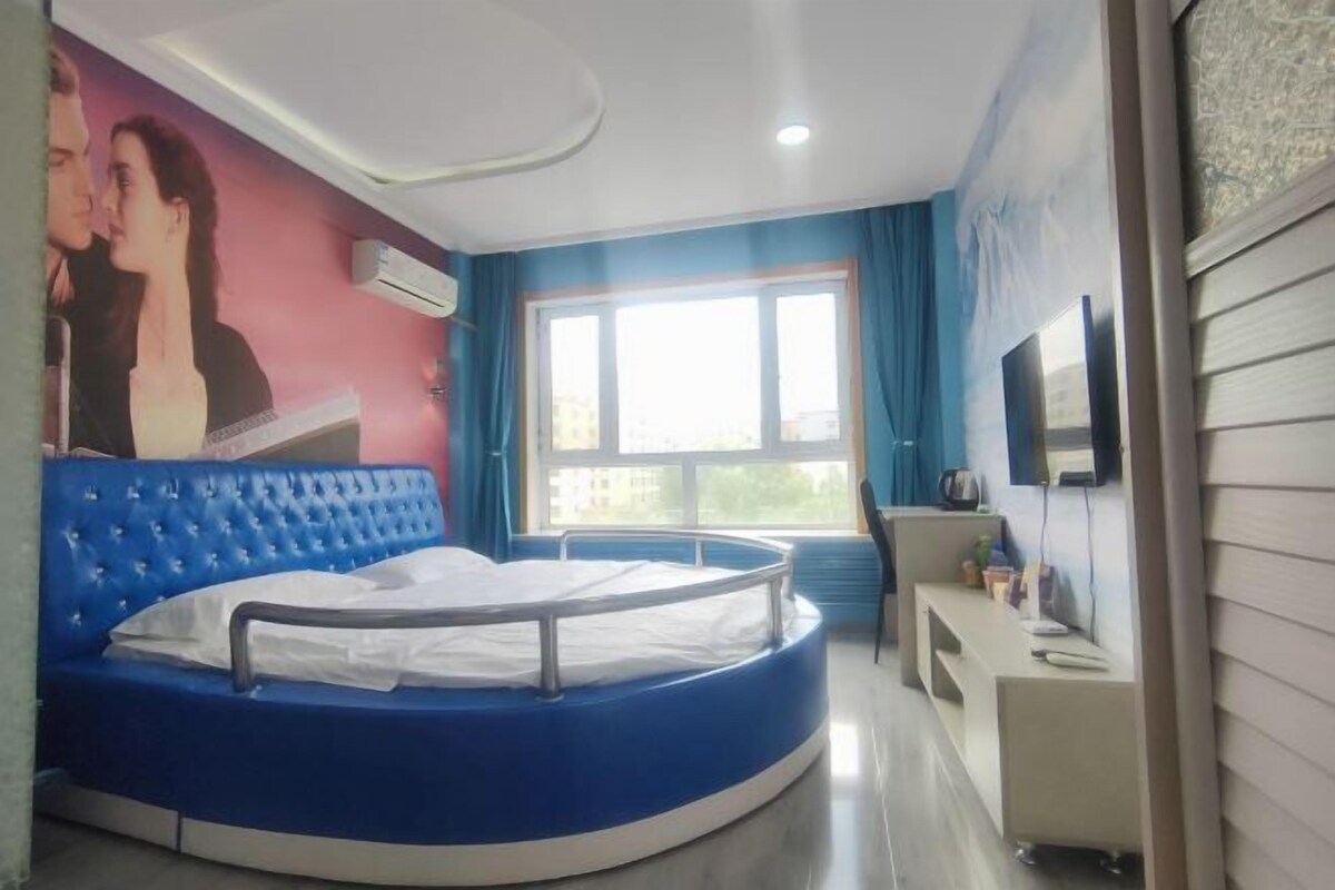 A circular bed with a white mattress and blue headboard in a room with a large window, a flat-screen TV, and a small desk.