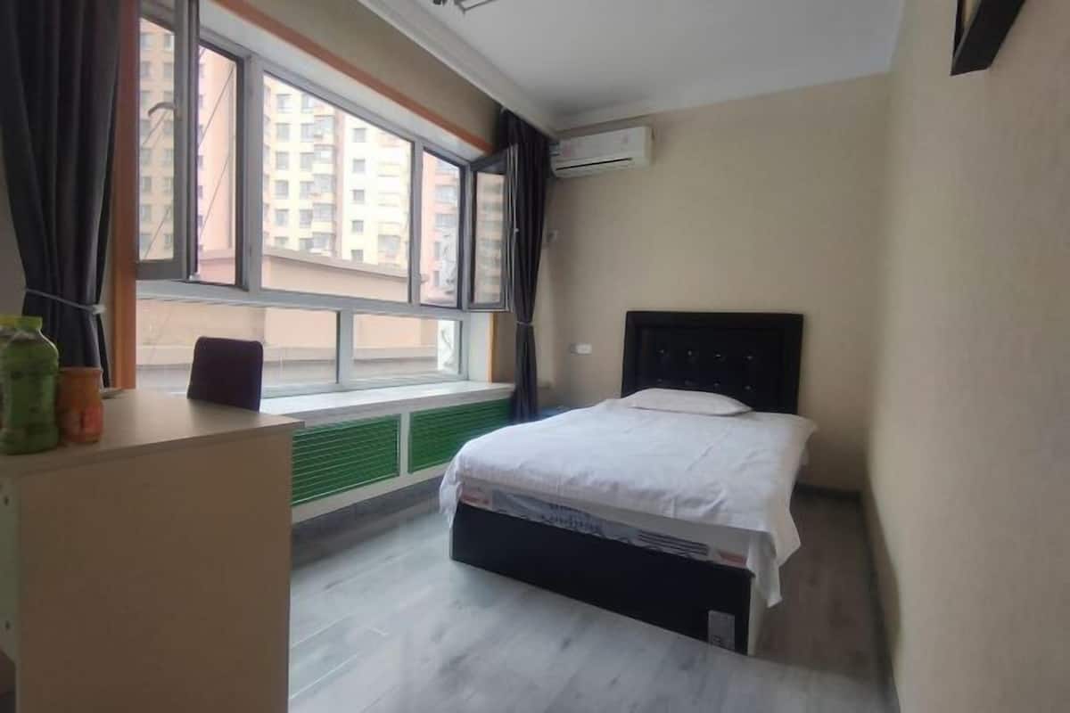 A bedroom with a bed, a window with curtains, a radiator, and a desk with a vase.