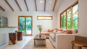A modern living room with a sofa, coffee table, and wooden side tables. There is a kitchen area with a refrigerator and a window with a view of greenery. - Unique beach side villa Rocamar Santa Teresa (Puntarenas Province)