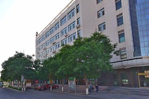 Un edificio de varios pisos con fachada moderna, una zona peatonal con árboles y automóviles estacionados.