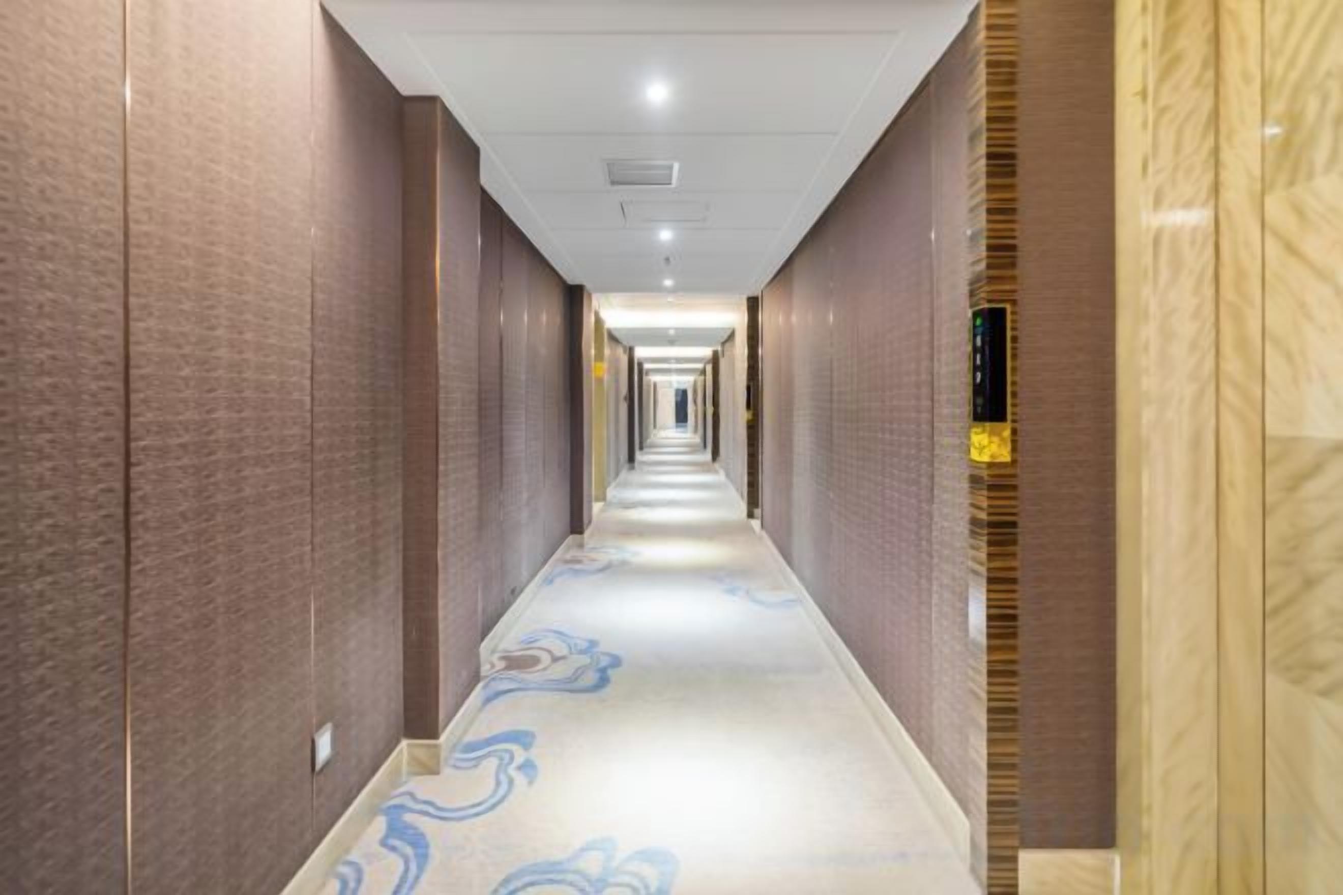 Un long couloir étroit, typique des hôtels, avec un tapis à motifs et un sol de couleur claire.