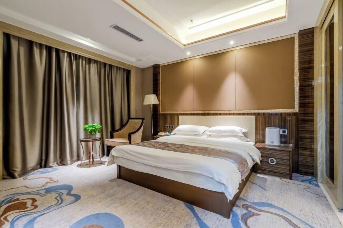 A hotel room with a large bed, a bedside table with a lamp, a small side table with a plant, and a large mirror on the wall.