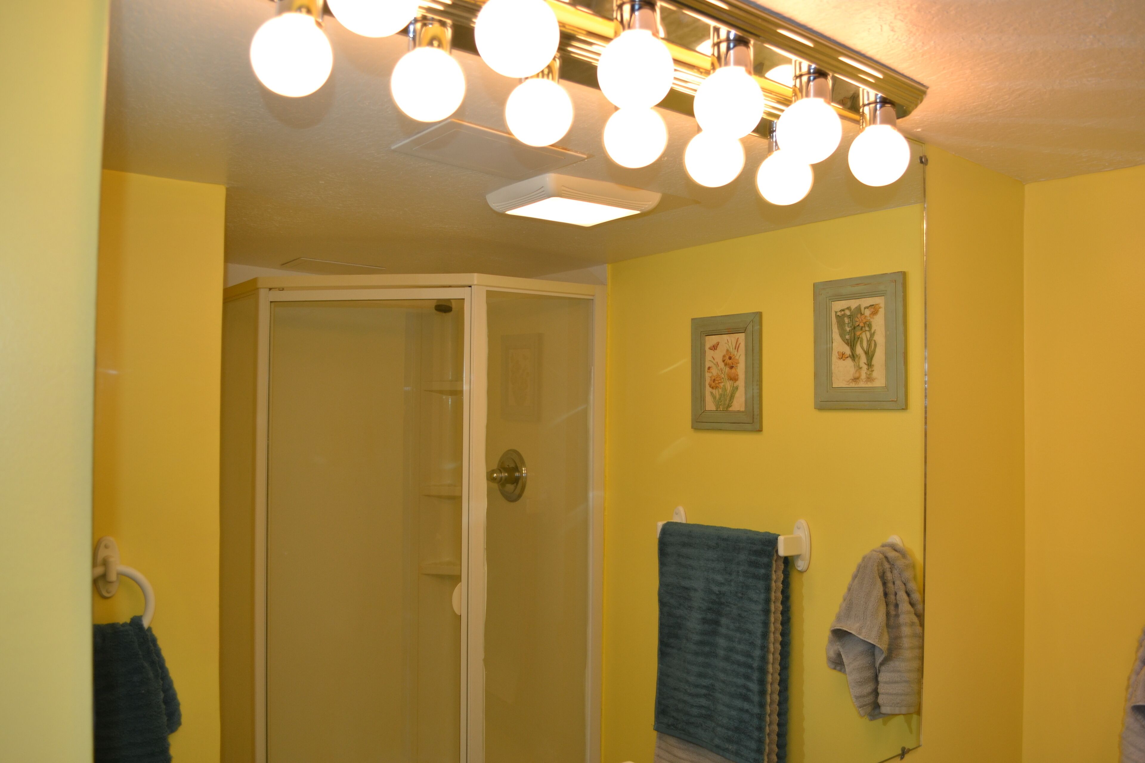 Une salle de bain aux murs jaunes, avec une porte de douche en verre et deux tableaux encadrés accrochés au mur.