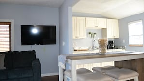 A living room with a dark grey couch, a flat-screen TV mounted on the wall, and a kitchen area with light wooden cabinets, a coffee maker, and a small plant. - Akron (Portage Lakes) Peaceful Lakefront Cottage with Kayaks  (Akron)