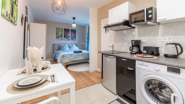 Cocina compacta con lavavajillas, microondas y cafetera. Se ve una cama con colcha azul estampada al fondo.