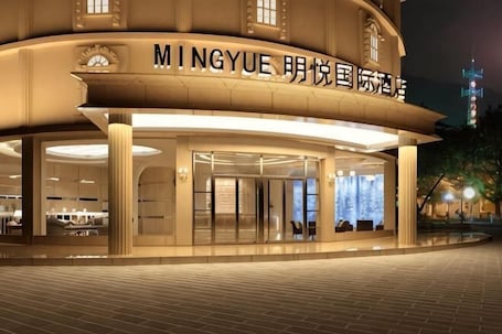 A modern hotel with a curved facade, large glass entrance, and signage in multiple languages.. Mingyue International Hotel