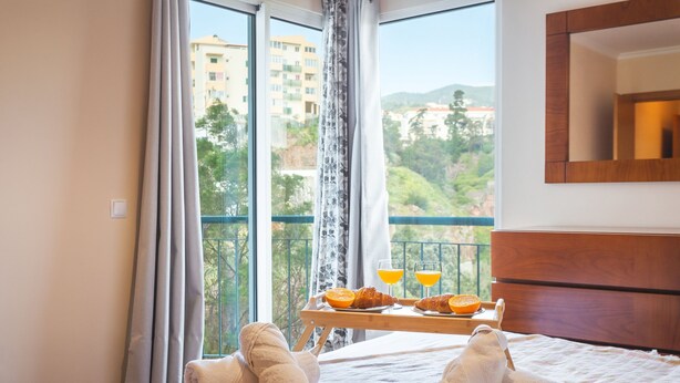 A hotel room with a bed, a tray with croissants and orange juice, and a view of buildings and trees through the window. - Charming Garajau by Atlantic Holiday (Santa Cruz)