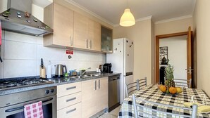 A modern kitchen with a gas stove, oven, and refrigerator, a dining table with a checkered tablecloth, and a hanging pendant light. - Charming Garajau by Atlantic Holiday (Santa Cruz)