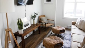 Ett vardagsrum med trägolv, en soffa, en fåtölj, ett bord med en växt, en lampa och en tv som är monterad på väggen.