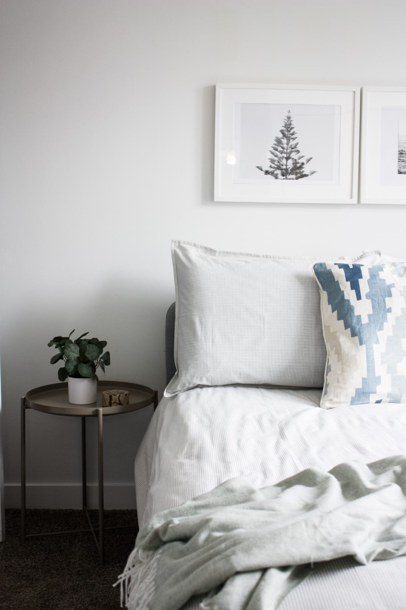 Une chambre à coucher bien rangée, avec un lit, une petite table de chevet ornée d’une plante et deux tableaux encadrés accrochés au mur.