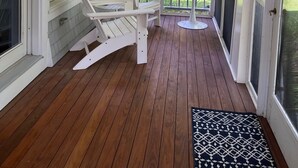 A covered porch with wooden flooring, two white Adirondack chairs, a small round table, and a view of a green lawn and trees. - Townhouse Cottage on Oceanfront Property Adj. Samoset Golf Resort+Lighthouse (Rockport)