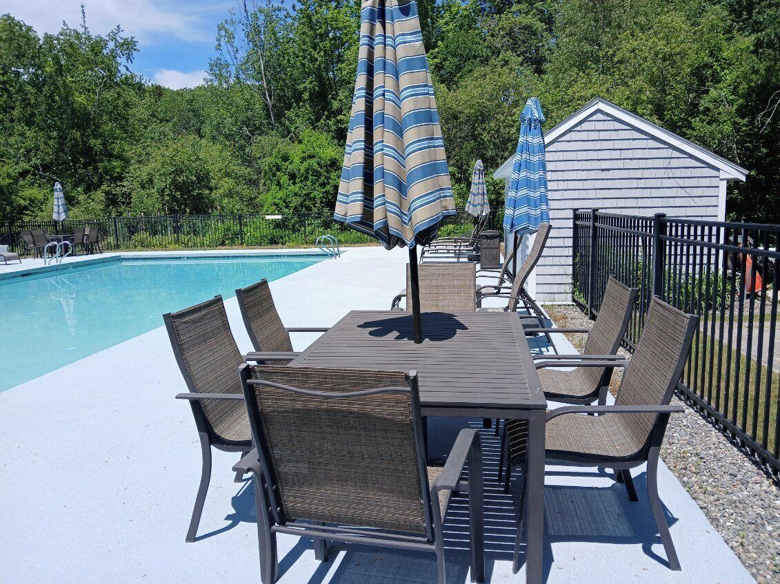 A poolside area with a table and chairs, umbrellas, and a small shed.