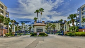 A gated entrance to a hotel with a sales office sign, palm trees, and a clear blue sky. - Pet Family Friendly condo 2 heated pools, hot tub, sauna, bocce Tennis, etc (New Smyrna Beach)