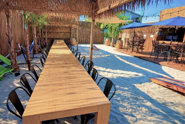 A long wooden table with chairs, a thatched roof, and a poolside area with umbrellas.