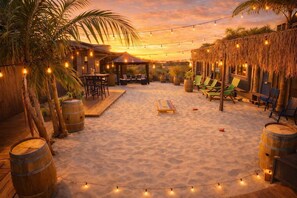 A beachside patio with wooden furniture, string lights, and palm trees.