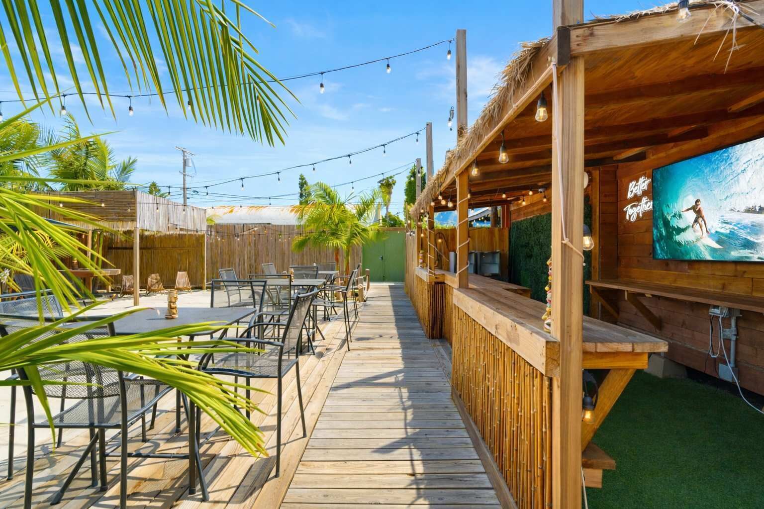 Una terraza de madera con mobiliario exterior y una zona de bar, rodeada de palmeras y un mural.