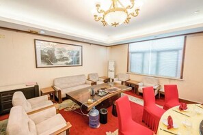 Hotellihuone, jossa on suuri ikkuna, kattokruunu, sohva, ruokapöytä punaisine tuoleineen sekä taulu seinällä.