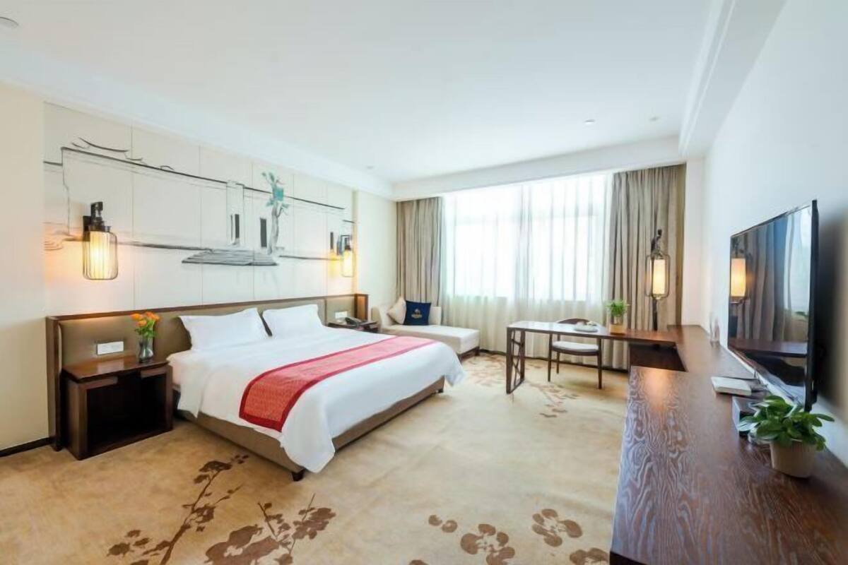 A hotel room with a bed, bedside tables, a desk, a chair, a television, and a large window with curtains.