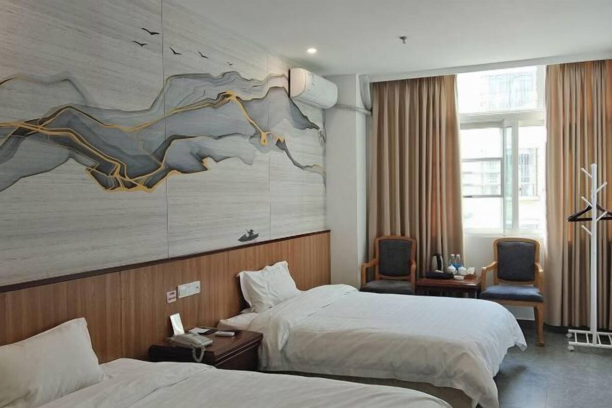 A hotel room with two beds, a wooden headboard, a wall-mounted artwork, a seating area with a table and chairs, and a window with curtains.