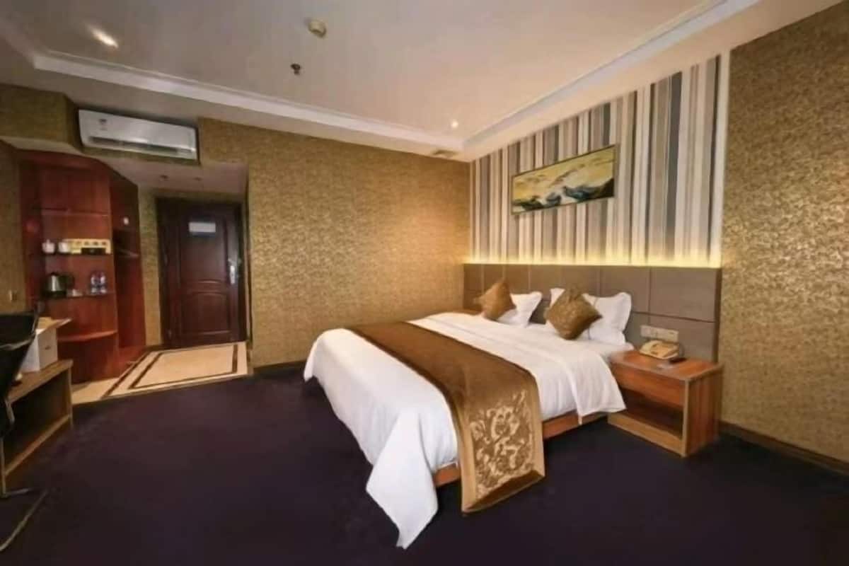 A hotel room with a bed, bedside table, nightstand, wardrobe, and an air conditioning unit.