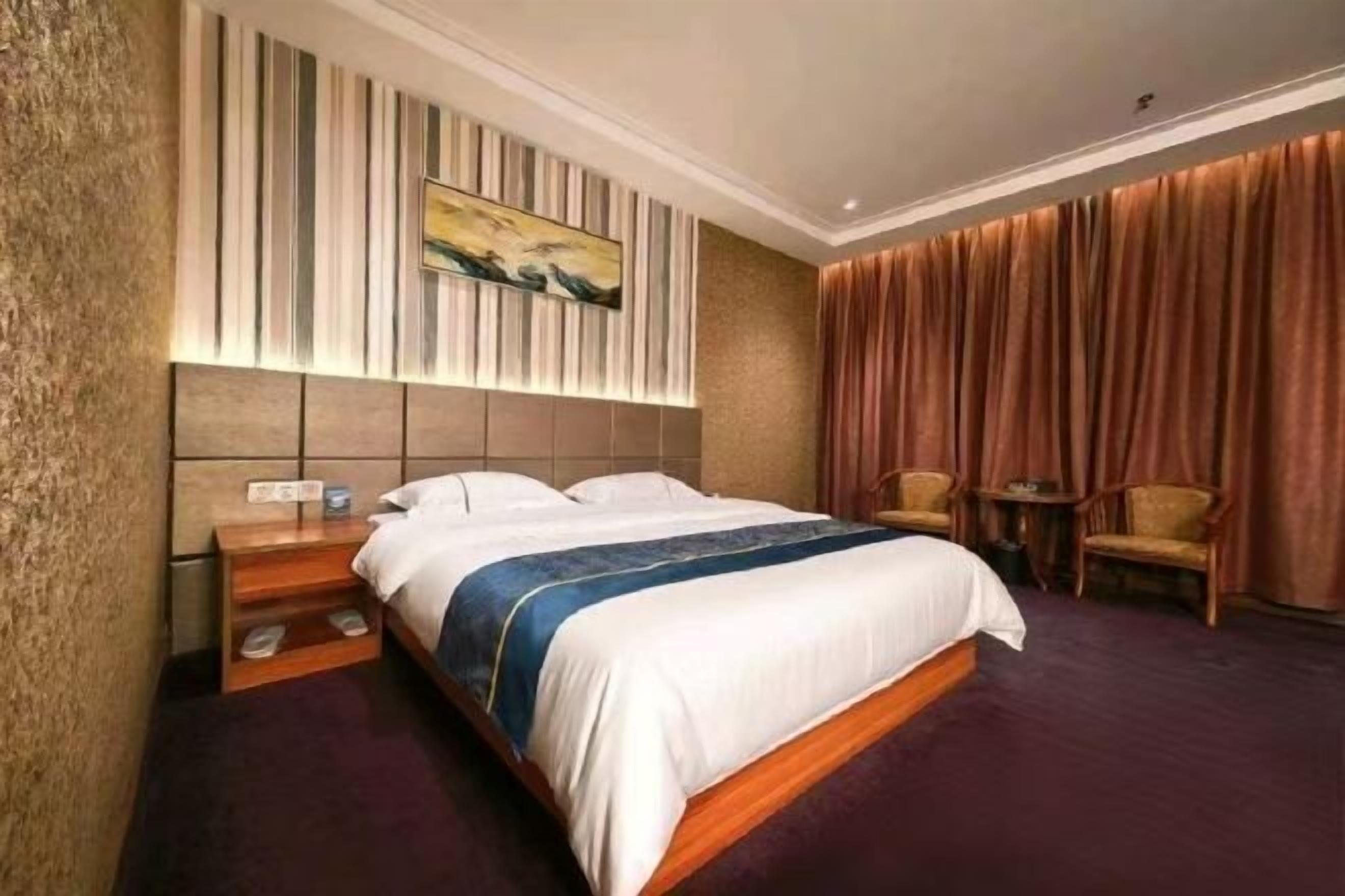 A hotel room with a bed, a nightstand, a chair, a small table, and a painting on the wall.