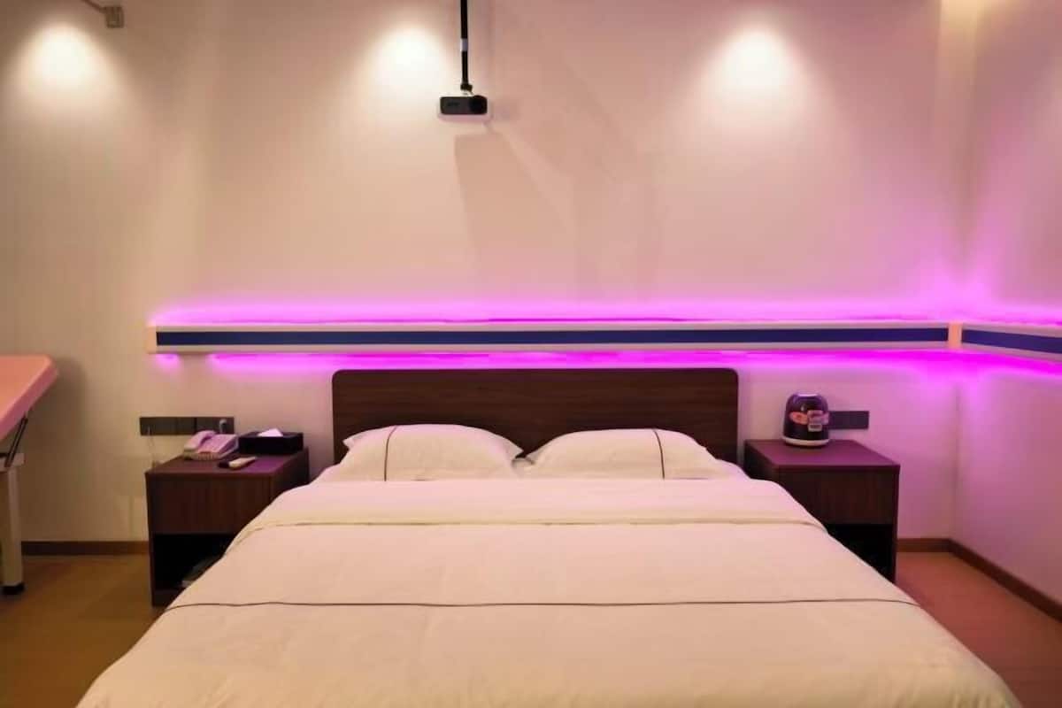 A hotel room with a bed, bedside tables, a nightstand, a lamp, and a wall-mounted shelf.