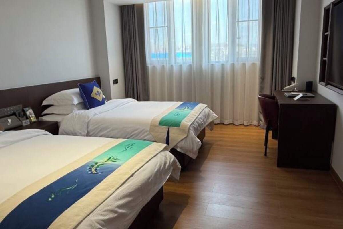 Habitación de hotel con dos camas, un escritorio, una silla y un amplio ventanal con cortinas.