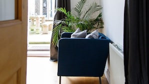 Un couloir étroit avec une chaise bleue, une grande plante en pot et une vue sur un espace extérieur aménagé avec une table et des chaises.