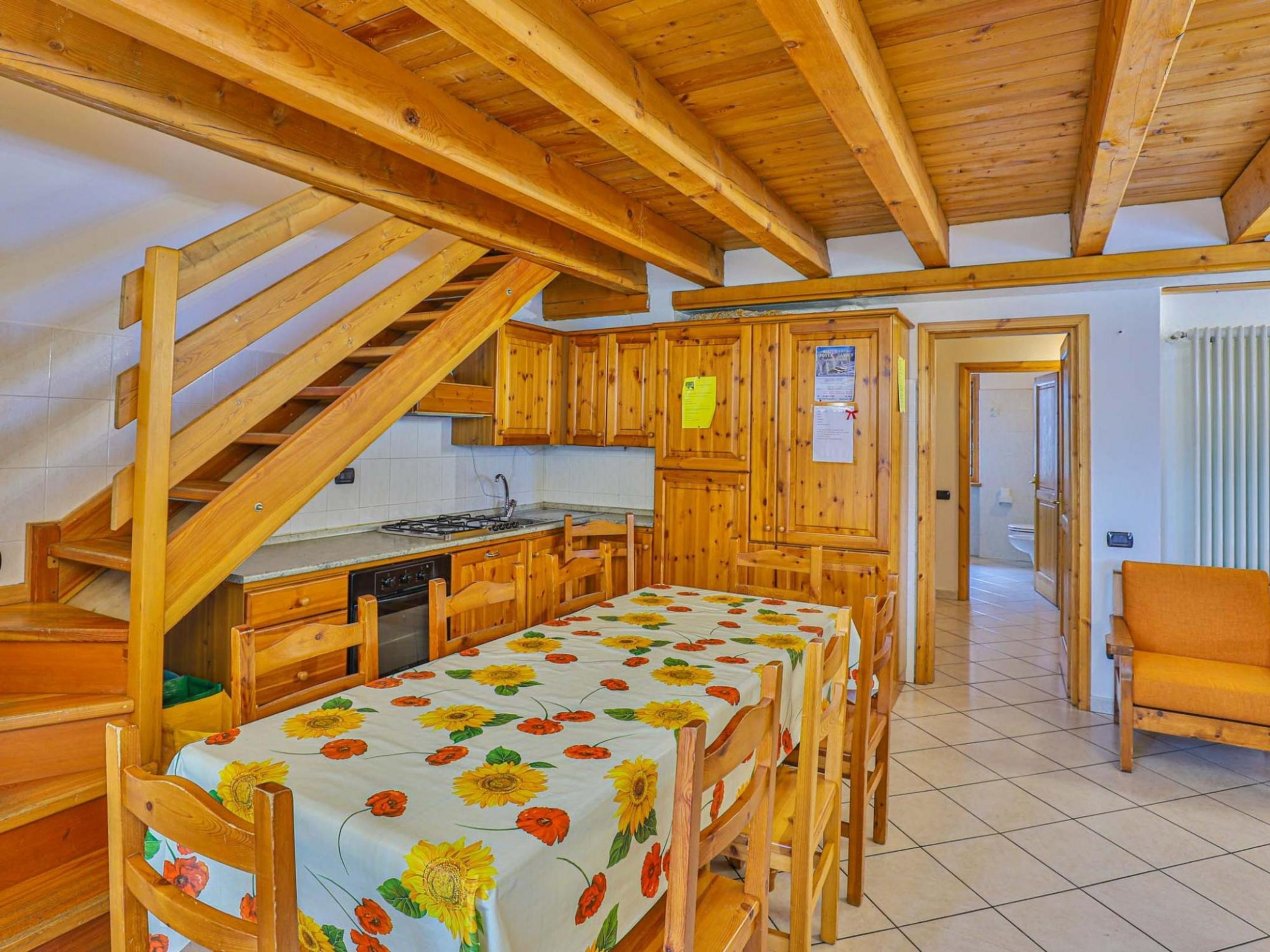 Eine rustikale Küche mit Holzsäulen, einem Esstisch mit Sonnenblumen-Tischdecke und einer Treppe, die in einen oberen Bereich führt.